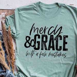 Puede incluir: Una camiseta azul claro con un gráfico negro que dice "mercy & grace with a few mistakes".