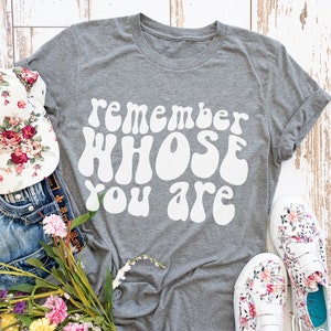May include: Gray t-shirt with the text "remember whose you are" in white, retro-style lettering. The shirt is laid out on a white wood background with a floral hat, denim shorts, and floral shoes.