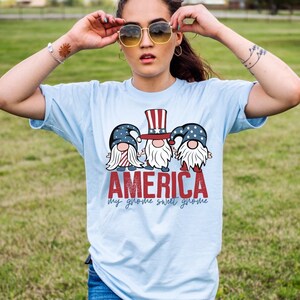 Puede incluir: Camiseta azul claro con un gráfico de tres gnomos con sombreros rojo, blanco y azul y el texto "AMERICA my gnome sweet gnome".