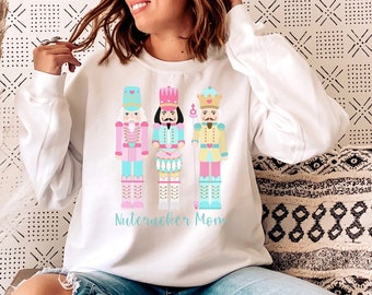 Puede incluir: Sudadera blanca con un gráfico de tres figuras de cascanueces y el texto "Nutcracker Mom".