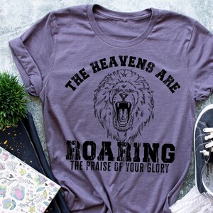Puede incluir: Una camiseta morada con un gráfico de león negro y el texto "The heavens are roaring the praise of your glory".