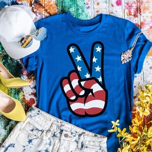 Puede incluir: Una camiseta azul con un gráfico de signo de paz con los colores de la bandera estadounidense. El signo de paz es rojo, blanco y azul. La camiseta está colocada sobre un fondo colorido con un sombrero blanco, zapatos amarillos y un par de gafas de sol.