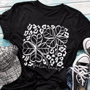 Pode incluir: Camiseta preta com um design de trevo branco e as palavras "feelin lucky". A camisa &eacute; combinada com um bon&eacute; com estampa em preto e branco, t&ecirc;nis pretos e shorts jeans em uma superf&iacute;cie de madeira azul.