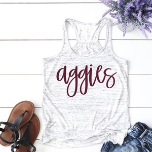 May include: White tank top with a maroon "Aggies" graphic. The tank top is on a white wooden surface with a pair of brown sandals and a pair of denim shorts.
