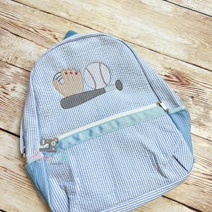 May include: A blue and white striped backpack with a baseball, bat, and glove embroidered on the front. The backpack has a mesh pocket on the side and a zippered pocket on the front.