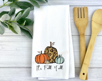 May include: White kitchen towel with a design of three pumpkins. The pumpkins are orange, teal, and leopard print. The text "It's Fall Y'all" is printed below the pumpkins.