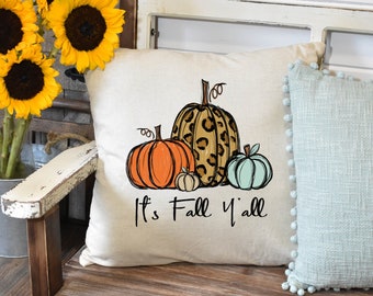 May include: A white throw pillow with a design of three pumpkins. The largest pumpkin is brown with a leopard print pattern. The middle pumpkin is orange and the smallest pumpkin is light blue. The text "It's Fall Y'all" is printed below the pumpkins.