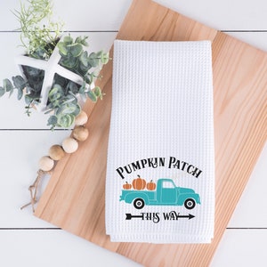 May include: White waffle-weave kitchen towel with a teal truck graphic carrying pumpkins. The text "PUMPKIN PATCH THIS WAY" is printed above and below the truck. The towel is on a wooden cutting board.