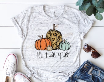 May include: A white t-shirt with a distressed look featuring a graphic of three pumpkins. The pumpkins are orange, green, and brown with a leopard print pattern. The text "It's Fall Y'all" is printed below the pumpkins.