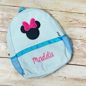 May include: A light blue and white striped backpack with a blue ribbon and a black and pink Minnie Mouse applique. The backpack is personalized with the name "maddie" in pink.