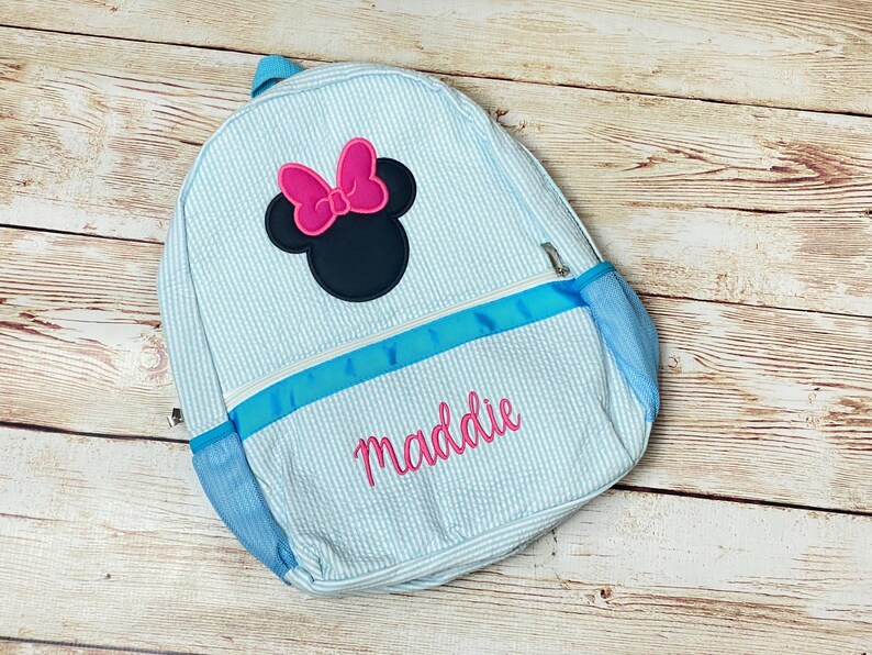 May include: A light blue and white striped backpack with a blue ribbon and a black and pink Minnie Mouse applique. The backpack is personalized with the name "maddie" in pink.