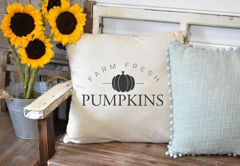 Puede incluir: Una almohada blanca con texto negro que dice "FARM FRESH PUMPKINS" y una silueta negra de una calabaza. La almohada est&aacute; sentada en un banco de madera con una almohada azul con pompones blancos.