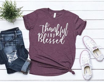 May include: A heathered maroon short-sleeve t-shirt with the white text "thankful AND Blessed" printed on the front, accompanied by a pair of dark blue distressed denim jeans and a pair of white low-top canvas sneakers.