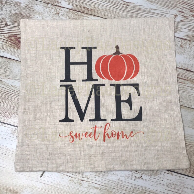 May include: A beige linen throw pillow with the words "HOME" in black letters, a red pumpkin graphic, and the text "sweet home" in cursive.