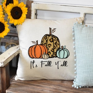 Puede incluir: Una almohada blanca con un dise&ntilde;o de cuatro calabazas. Las calabazas son naranja, marr&oacute;n con estampado de leopardo y azul claro. El texto "It's Fall Y'all" est&aacute; impreso debajo de las calabazas.