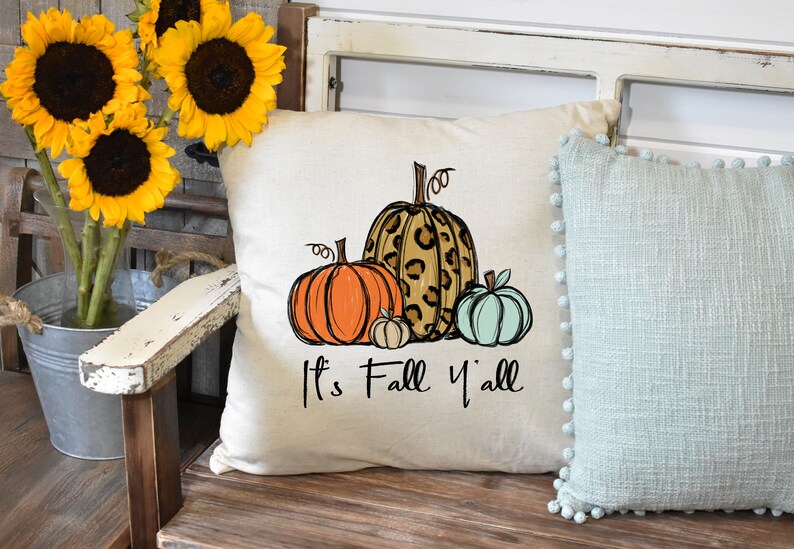 Puede incluir: Una almohada blanca con un dise&ntilde;o de cuatro calabazas. Las calabazas son naranja, marr&oacute;n con estampado de leopardo y azul claro. El texto "It's Fall Y'all" est&aacute; impreso debajo de las calabazas.
