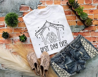 May include: A white short-sleeved t-shirt featuring a black line art graphic of a nativity scene under a star, with the text "BEST. DAY. EVER." printed below. The design depicts figures in a stable setting with surrounding stars.