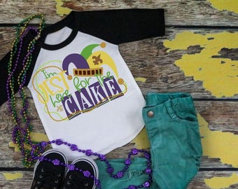 May include: A white raglan shirt with black sleeves featuring a graphic of a jester hat and the text I'm JEST for the CAKE, paired with teal pants, black sneakers, and a long strand of purple, green, and gold Mardi Gras beads.