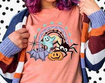 May include: A peach-colored t-shirt featuring a graphic print of a light blue ghost, a black spider, a purple bat, and an orange pumpkin centered under a rainbow. The shirt is worn with a long-sleeved, multi-colored striped cardigan.