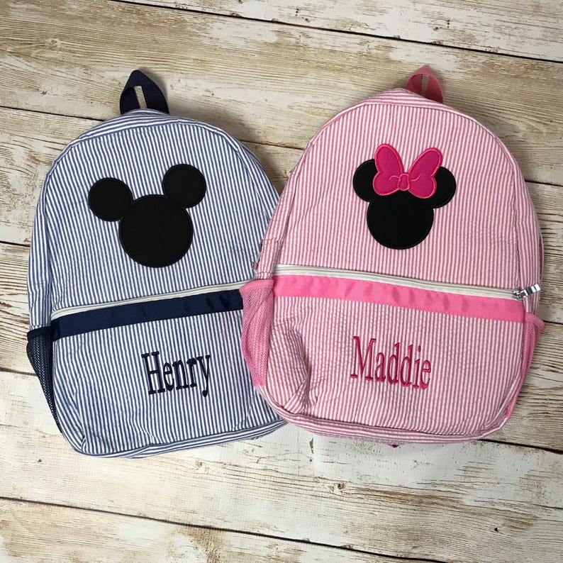 May include: Two personalized children's backpacks with Mickey Mouse and Minnie Mouse embroidery. The blue backpack has the name "Henry" embroidered on it. The pink backpack has the name "Maddie" embroidered on it.
