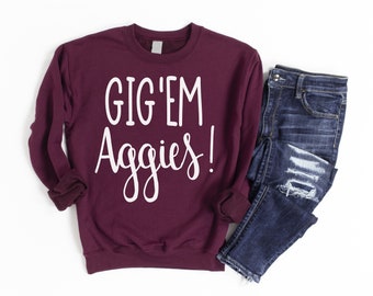 May include: Burgundy sweatshirt with white text that reads "Gig 'Em Aggies!"  The sweatshirt is paired with blue jeans.