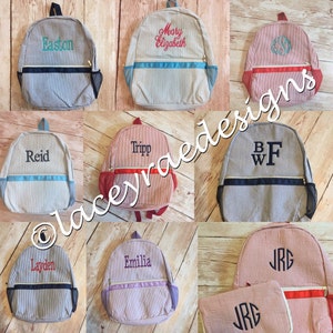May include: Nine personalized striped canvas backpacks with various colors and embroidered names and initials. The backpacks have a zippered pocket on the front and a mesh pocket on the side.