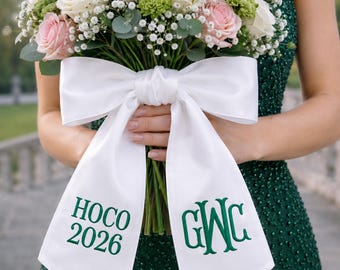 Puede incluir: Un ramo de flores con rosas de color rosa claro y blanco, flores de relleno verdes y hojas de eucalipto, atado con una cinta de satén blanca ancha. La cinta está bordada con texto verde oscuro que dice HOCO 2026 y el monograma GWC.