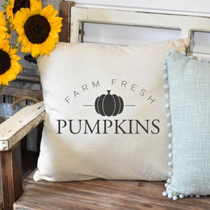 Puede incluir: Una almohada blanca con texto negro que dice "Farm Fresh Pumpkins" y una silueta negra de una calabaza. La almohada est&aacute; sentada en un banco de madera con una almohada azul claro con pompones blancos en el borde.