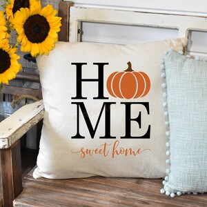 May include: A white throw pillow with the word "HOME" in black letters, a pumpkin graphic, and the text "sweet home" in cursive. The pillow is on a wooden bench with a blue throw pillow next to it. There are sunflowers in a metal bucket in the background.