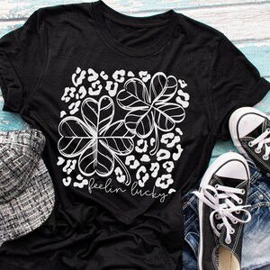 Pode incluir: Camiseta preta com um design de trevo branco e o texto "feelin lucky". A imagem tamb&eacute;m mostra um bon&eacute; de tweed e t&ecirc;nis de lona pretos. O design inclui um fundo com estampa de leopardo.