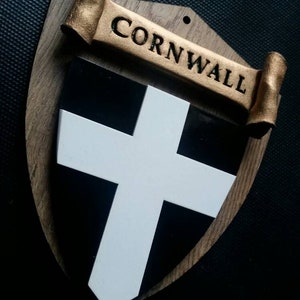 May include: A wooden shield with a white cross on a black background. The shield is decorated with a gold scroll with the word "CORNWALL" written on it.