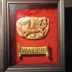 Puede incluir: Escultura en relieve enmarcada con un diseño detallado de dragón sobre una pancarta que dice "WALES". La obra de arte está sobre un fondo rojo dentro de un marco negro, creando un contraste llamativo.