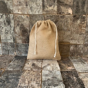 Pode incluir: Uma bolsa de juta bege com fecho de cord&atilde;o branco. A bolsa &eacute; feita de um material grosseiro e tecido e est&aacute; colocada contra um fundo de madeira envelhecida.