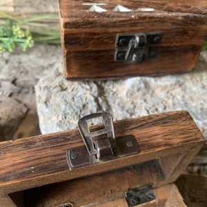 May include: A wooden box with a hinged lid and a metal latch. The box is made of dark wood with a natural finish. The latch is silver and has a simple design.