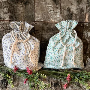 May include: Two fabric drawstring pouches featuring a white base with small floral patterns. The left pouch has blue and purple flower prints, while the right pouch has teal and green flower prints. Both bags include white rope drawstrings.
