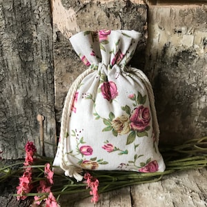 May include: A small white drawstring bag with a floral pattern featuring pink and yellow roses. The bag is made of fabric and has a white drawstring.