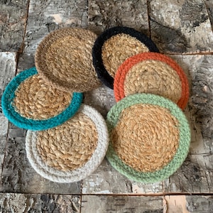 May include: A selection of circular coasters crafted from woven natural fibres. Each coaster features a different coloured border, such as teal, white, green, orange, black, and brown. The coasters are displayed on a rustic wooden surface.