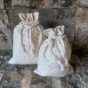 May include: Two off-white fabric drawstring pouches featuring a repeating pattern of small red stars. Each bag is cinched at the top with a thick white braided rope cord that ties in the front, creating a gathered ruffle effect at the opening.