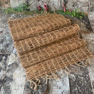 May include: A woven brown table runner made from natural fibres with frayed edges. The runner is folded in thirds and has a rustic, natural look.