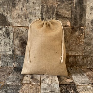 Pode incluir: Uma bolsa bege de juta com fecho de cord&atilde;o branco. A bolsa &eacute; feita de um material grosso e tecido e est&aacute; cheia. O fundo &eacute; uma parede com pain&eacute;is de madeira.