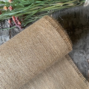 Puede incluir: Un rollo de tela de arpillera marr&oacute;n natural con un borde deshilachado. La tela est&aacute; tejida con un tejido apretado y uniforme.