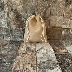 Pode incluir: Uma pequena bolsa de juta bege com fecho de cord&atilde;o branco. A bolsa &eacute; feita de um tecido grosso e tecido e est&aacute; sobre um fundo de madeira.