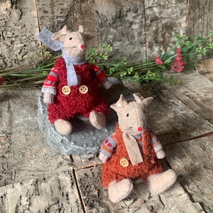 May include: Two handmade goat plush toys. One goat is wearing a red outfit with a grey scarf and the other is wearing an orange outfit with a beige scarf. Both have button closures and are sitting on a grey rock.