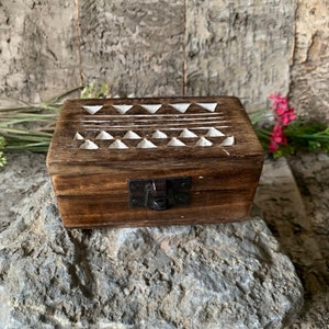 May include: A small wooden box with a hinged lid and a latch. The lid is decorated with a white geometric pattern of triangles and lines. The box is sitting on a rock.