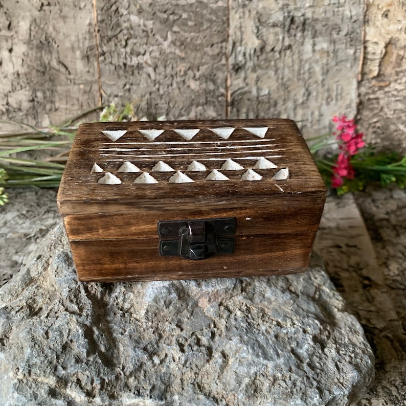 May include: A small wooden box with a hinged lid and a latch. The lid is decorated with a white geometric pattern of triangles and lines. The box is sitting on a rock.