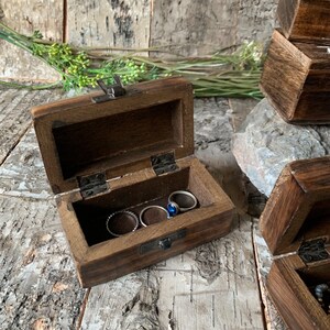 May include: A small wooden box with a hinged lid, open to reveal a compartment with four rings inside. The box is made of dark brown wood and has a metal latch.