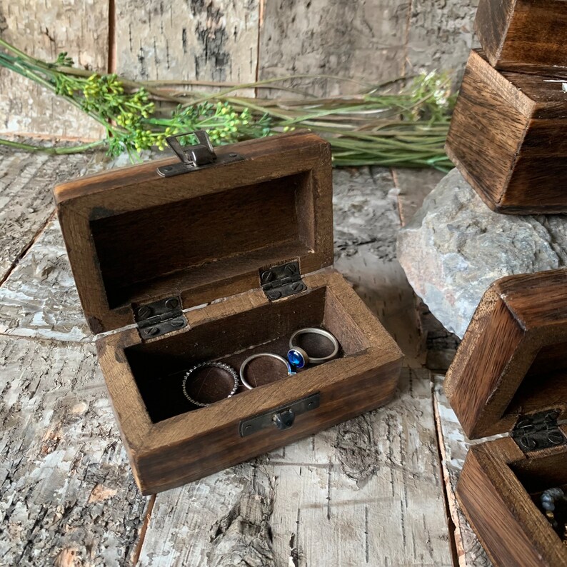 May include: A small wooden box with a hinged lid, open to reveal a compartment with four rings inside. The box is made of dark brown wood and has a metal latch.