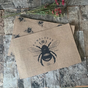 May include: Two brown hessian placemats with a black bee design. The top placemat has a bee with the text "Bumblebee Trade Mark" around it. The bottom placemat has a bee design in the corner.