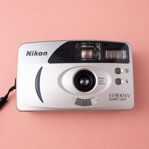 May include: A silver Nikon EF500SV Quartz Date 35mm film camera with a black strap. The camera has a black lens with the text "Nikon Lens 28mm" printed on it.