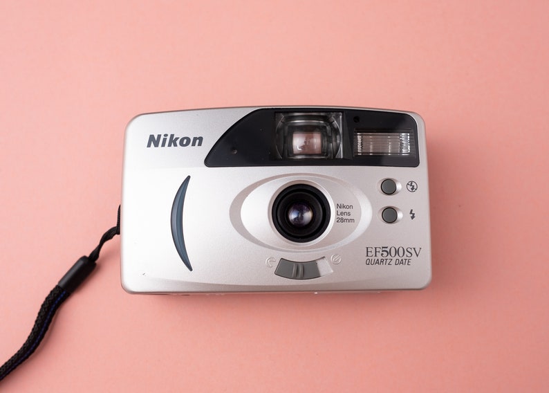 May include: A silver Nikon EF500SV Quartz Date 35mm film camera with a black strap. The camera has a black lens with the text "Nikon Lens 28mm" printed on it.